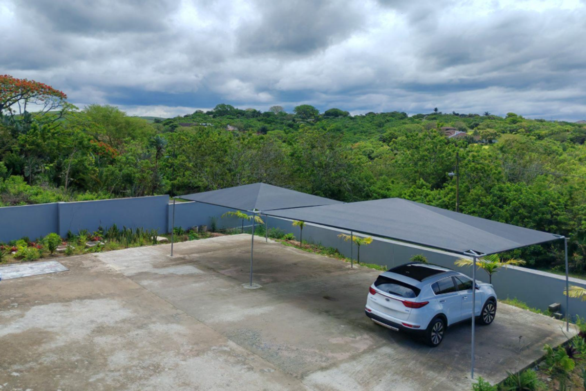 An outdoor parking area featuring a large shade sail structure, concrete paving, and perimeter boundary walls overlooking a lush green landscape.