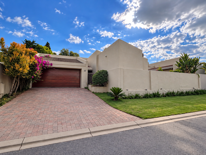 Front elevation of a townhouse featuring a paved driveway, timber garage door, and well-maintained boundary wall with established landscaping.