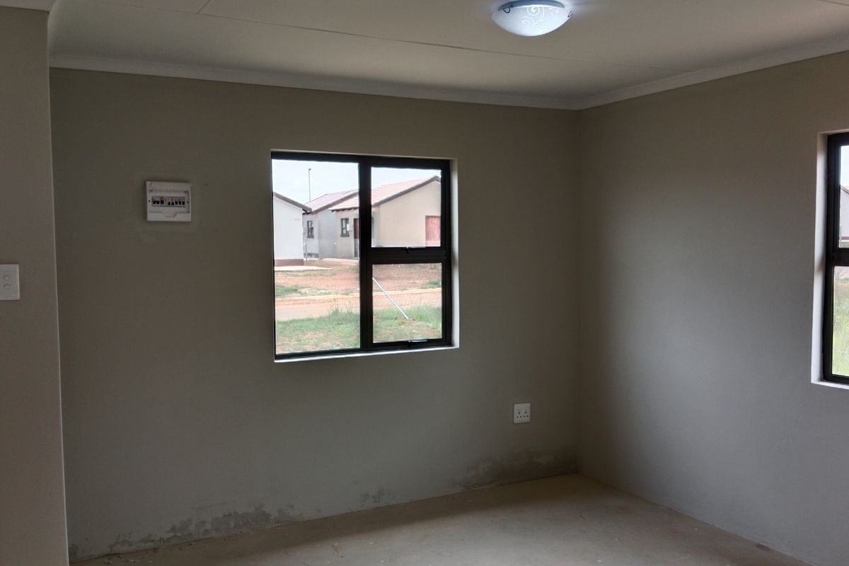 An empty, neutral-toned living area featuring aluminium window frames, an installed distribution board, and basic ceiling lighting, requiring minor finishing touches.