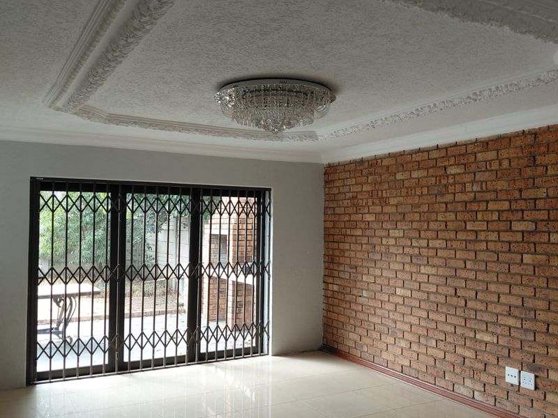 Spacious living room featuring an exposed brick wall, decorative ceiling cornice with a crystal chandelier, and tiled flooring leading to a secure patio.