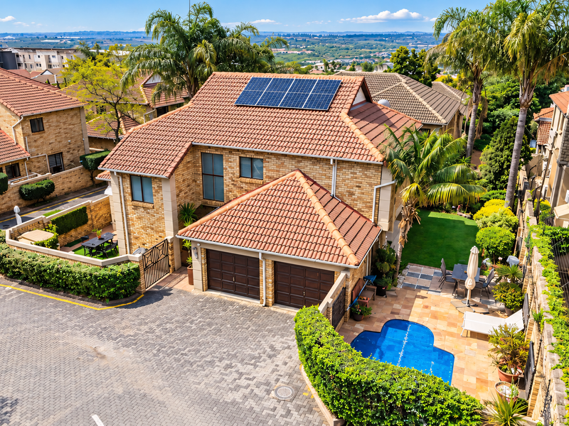 Aerial view of a townhouse featuring a tiled roof with solar panels, a double garage, a private swimming pool, and a landscaped garden.