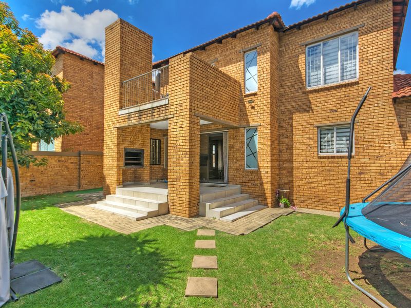 Rear view of a double-story brick townhouse featuring a covered patio with a built-in braai, an upper-level balcony, and a private garden area.