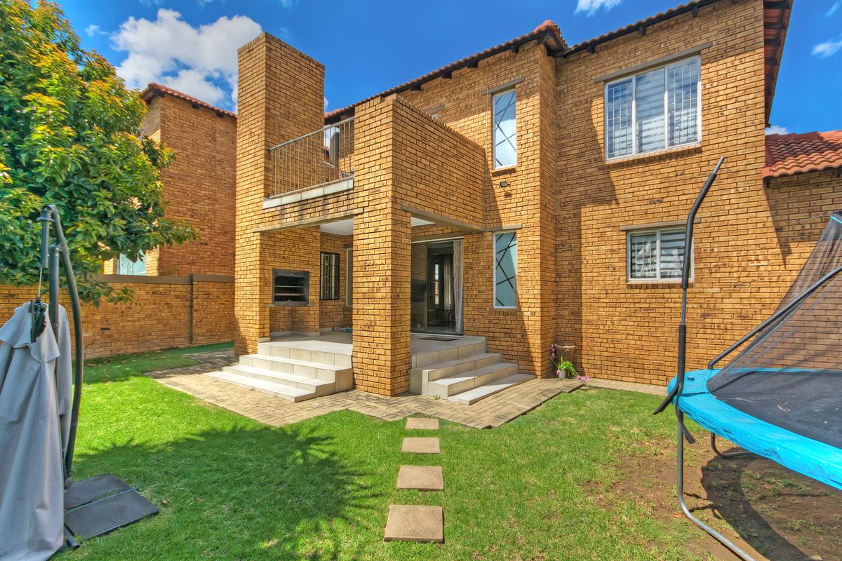 Rear view of a double-story brick townhouse featuring a covered patio with a built-in braai, an upper-level balcony, and a private garden area.
