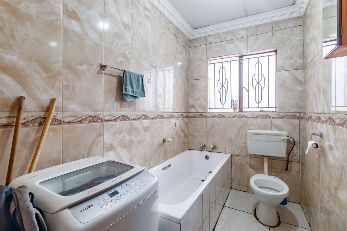 A functional bathroom featuring full-height marble-effect wall tiling, a standard bathtub, and a toilet, with security bars fitted to the window.