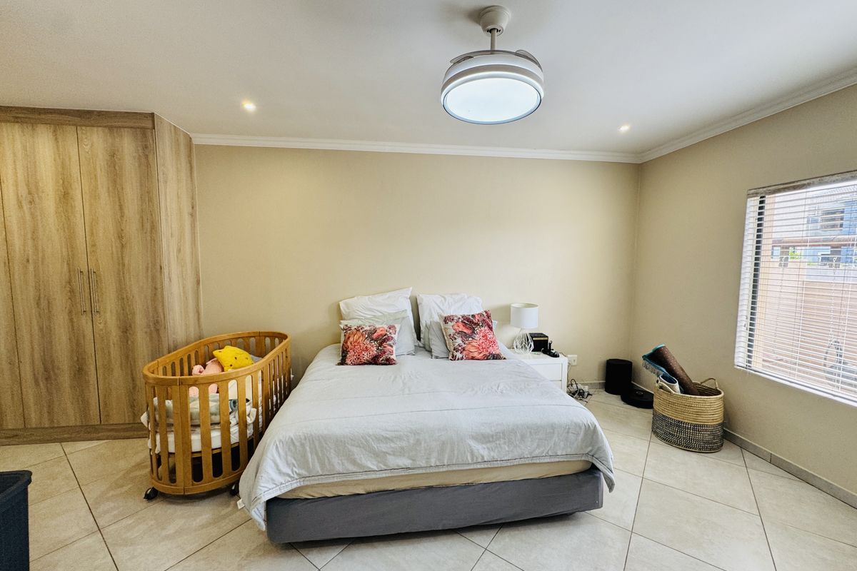 A spacious bedroom featuring neutral tiled flooring, built-in wooden cupboards, a modern ceiling fan, and large windows with venetian blinds.