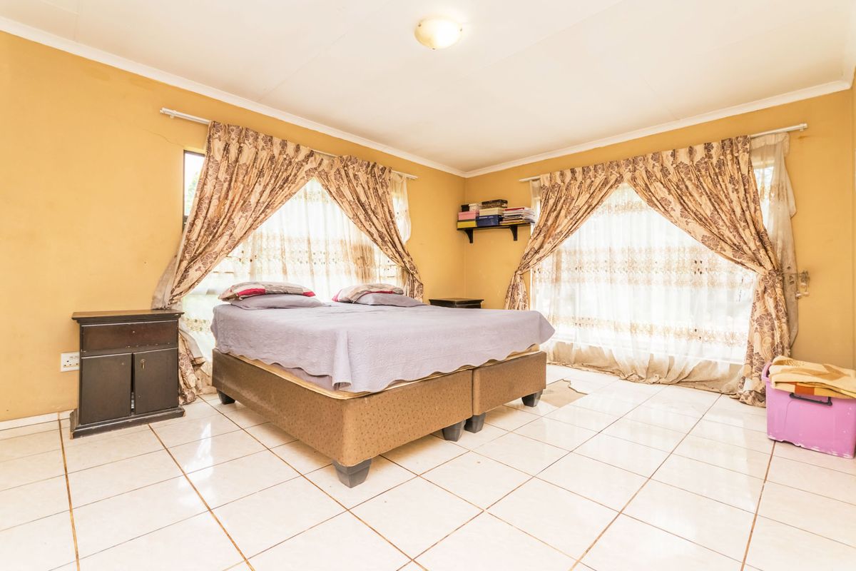 A bedroom featuring tiled flooring and dual windows with curtains. The room shows signs of wear, including cracked wall plaster and dated paint finishes.