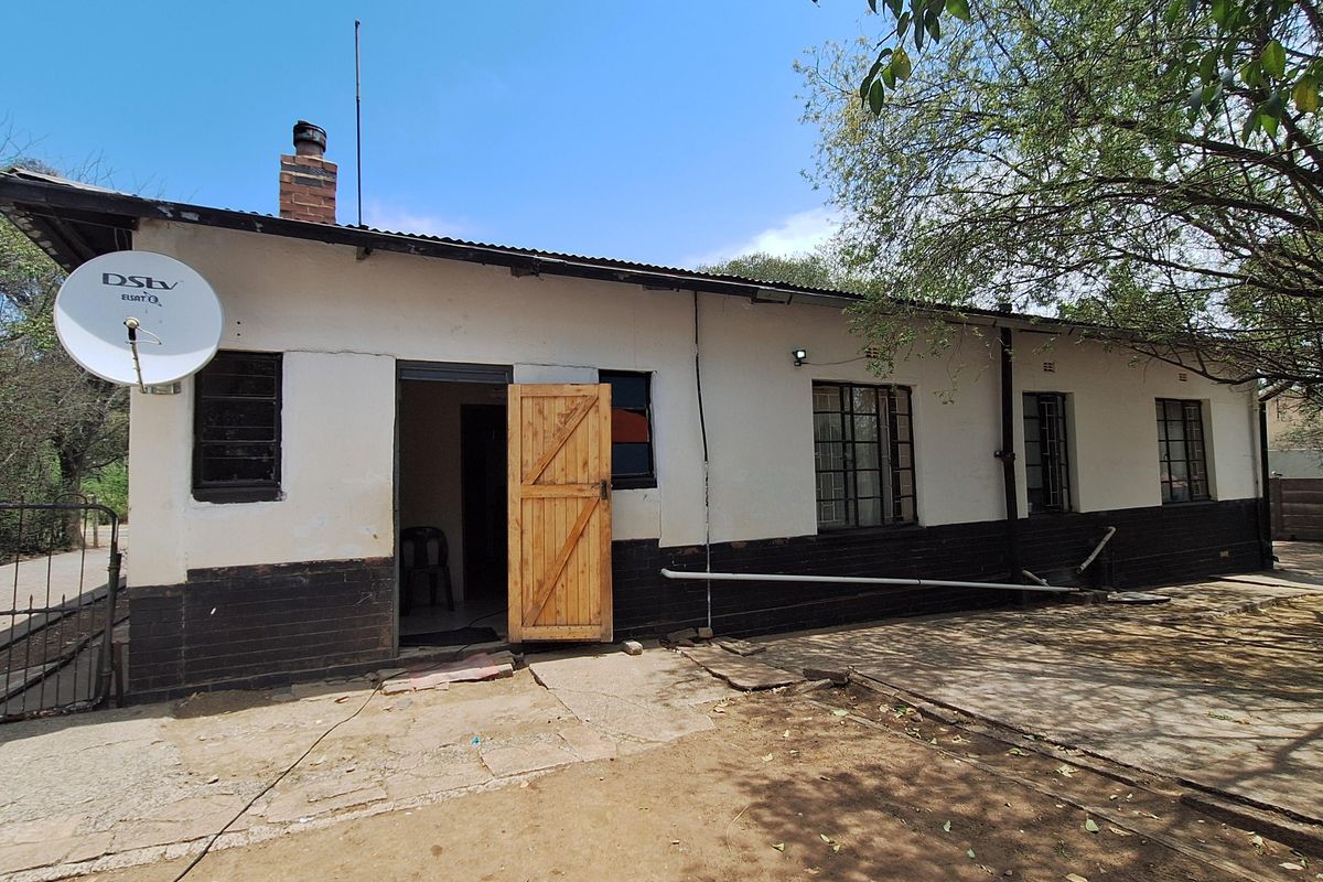 Rear exterior view of a single-story house featuring a brick chimney, security bars on windows, and a wooden entrance door.