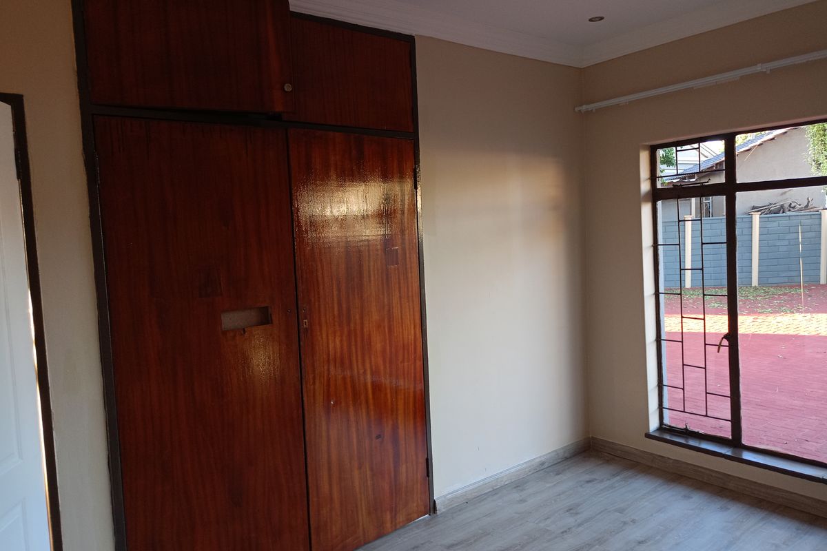 A bedroom featuring built-in wooden cupboards, laminate flooring, and a window with security bars overlooking the exterior area.
