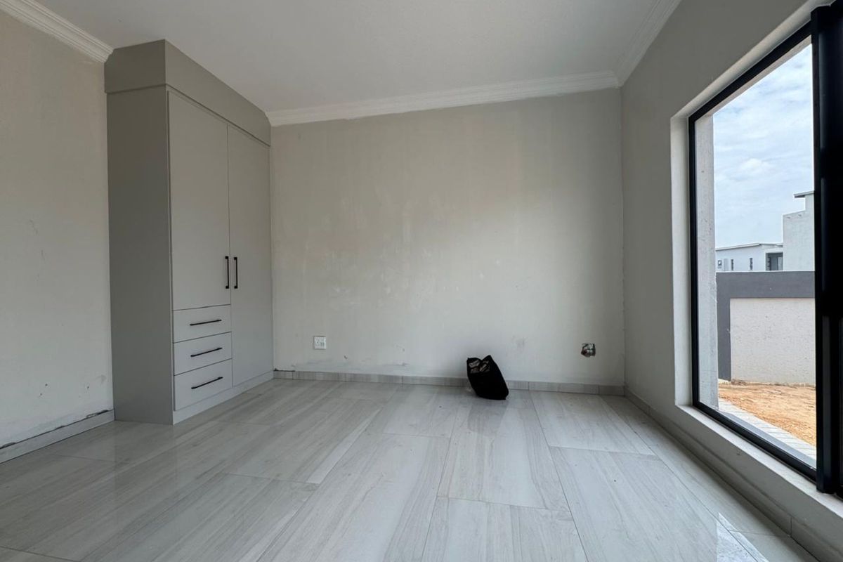 A bright, empty bedroom featuring modern light-toned floor tiles, built-in cabinetry, and a large window providing natural light.