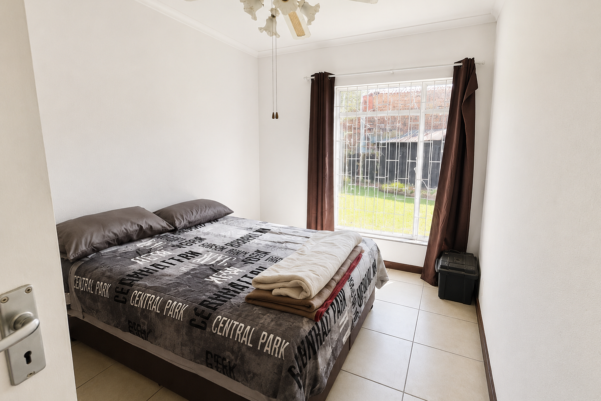 A functional bedroom featuring tiled flooring, a ceiling fan, and a window fitted with security bars, offering a view of the rear garden area.