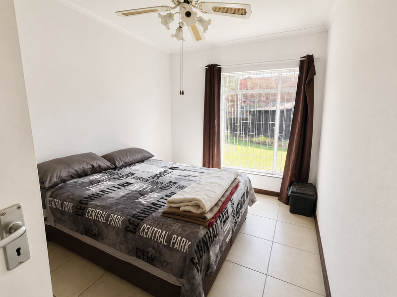 A functional bedroom featuring tiled flooring, a ceiling fan, and a window fitted with security bars, offering a view of the rear garden area.