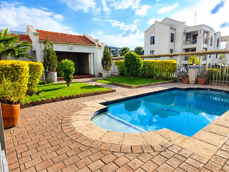 A well-maintained communal pool area featuring a paved deck, landscaped garden, and a covered outdoor space within an apartment complex.