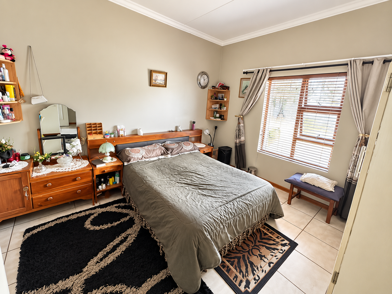 A well-lit bedroom featuring tiled flooring, a built-in headboard, and large windows with venetian blinds, offering a functional and comfortable living space.