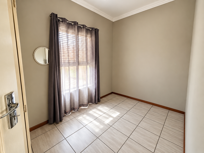 A clean, empty bedroom featuring neutral wall tones, tiled flooring, crown molding, and a window fitted with blinds and curtains.