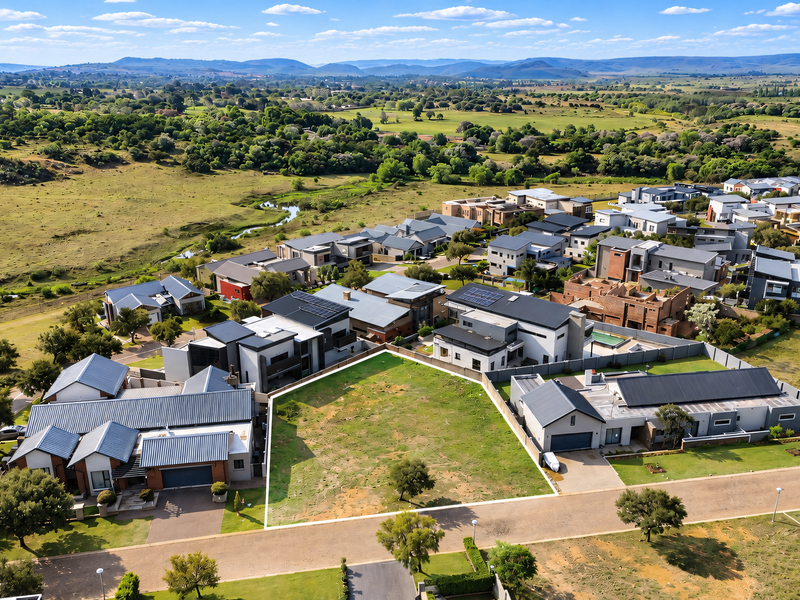 An aerial view of a vacant residential plot situated within a developed estate, featuring surrounding homes and scenic natural landscape views.