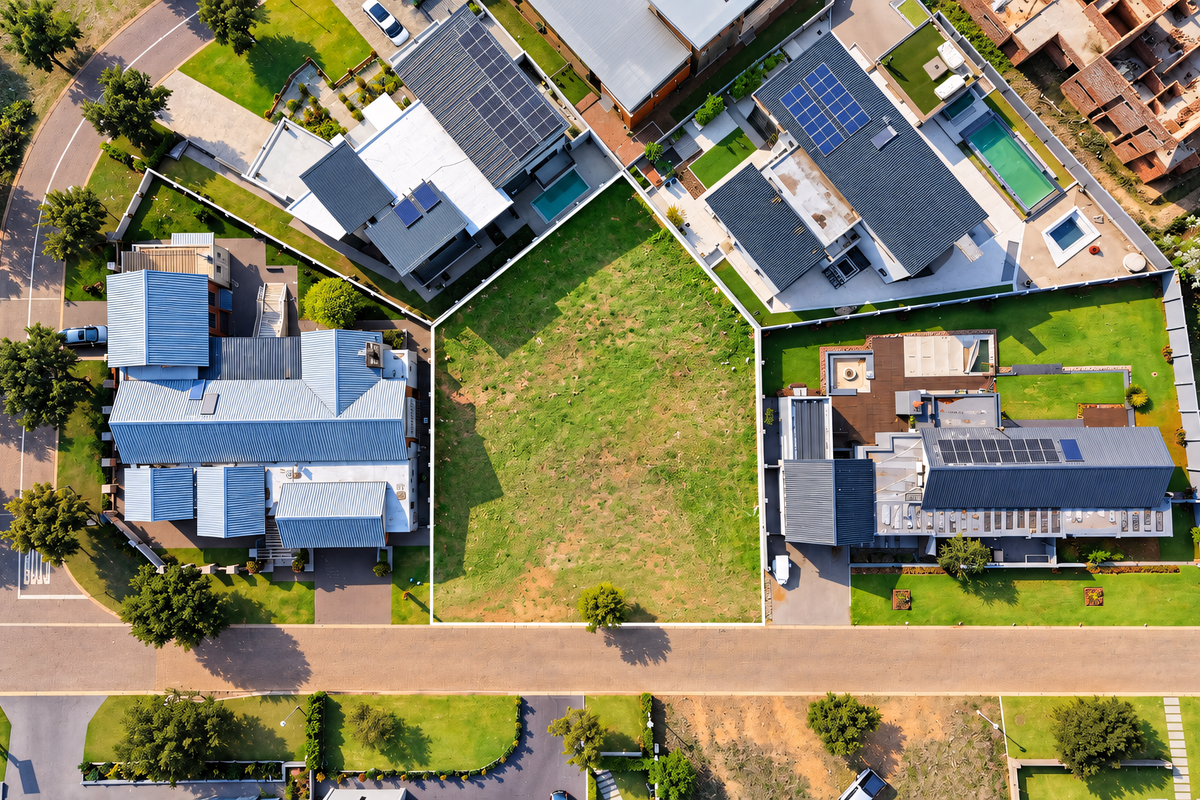 An aerial view of a vacant residential plot situated between established modern homes in a developed estate, featuring clear boundary lines and street access.