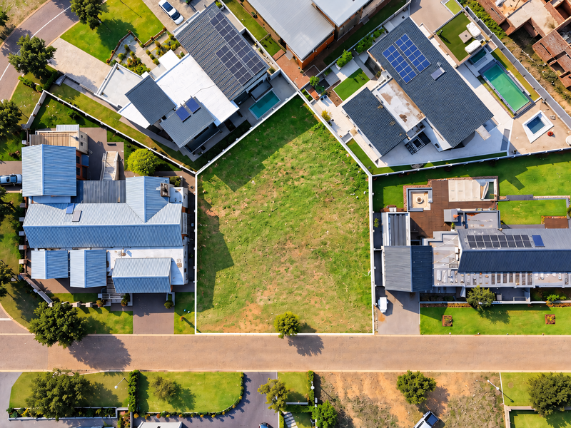 An aerial view of a vacant residential plot situated between established modern homes in a developed estate, featuring clear boundary lines and street access.