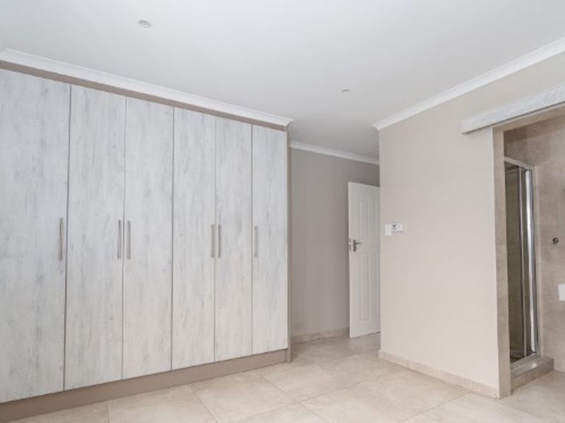 Spacious bedroom featuring extensive built-in cupboards, neutral tiled flooring, and a convenient en-suite bathroom with a shower and wall-mounted basin.
