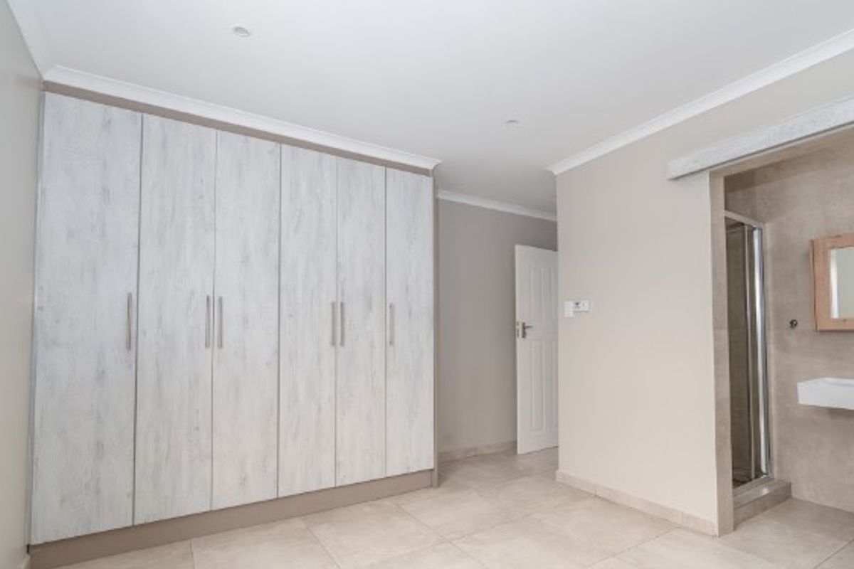 Spacious bedroom featuring extensive built-in cupboards, neutral tiled flooring, and a convenient en-suite bathroom with a shower and wall-mounted basin.