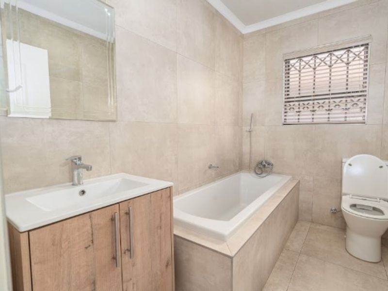 A modern, neutral-toned bathroom featuring a built-in bathtub, vanity cabinet with basin, and a toilet, finished with floor-to-ceiling wall tiles.