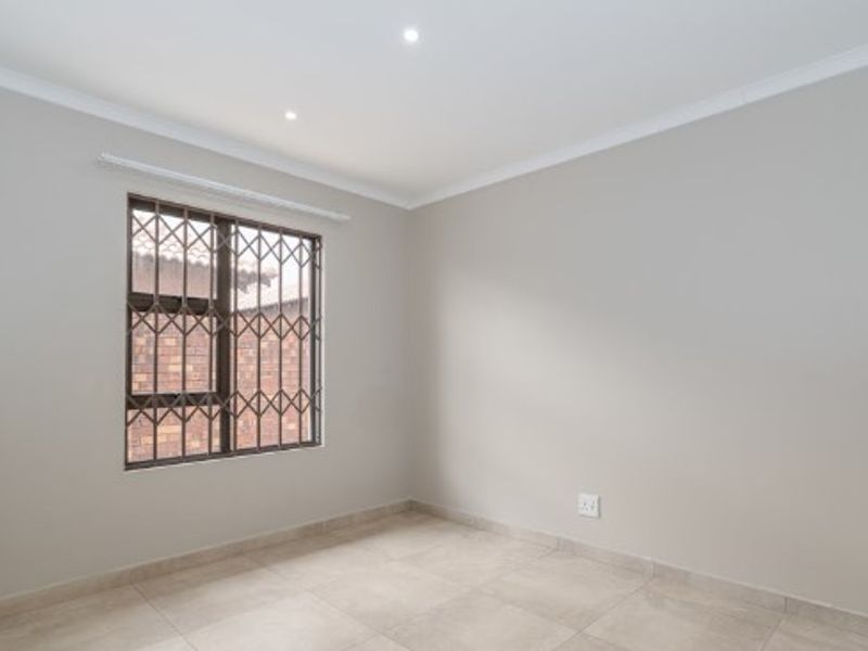 A clean, empty bedroom featuring neutral tiled flooring, recessed downlights, and a window fitted with a security gate.