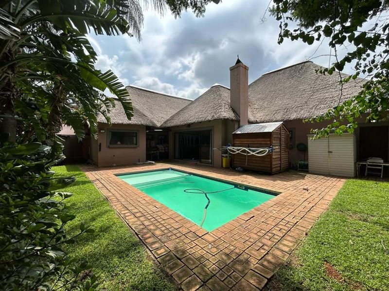 Rear garden area featuring a swimming pool, brick paved surround, and a traditional thatch-roofed house with a prominent chimney.