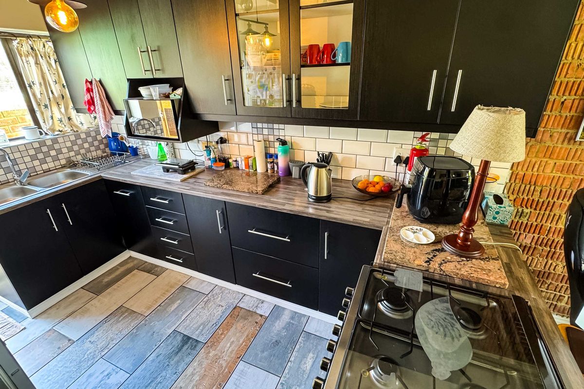 A functional kitchen featuring dark cabinetry, wood-look laminate countertops, a stainless steel double sink, and a built-in gas hob with an exposed brick feature wall.
