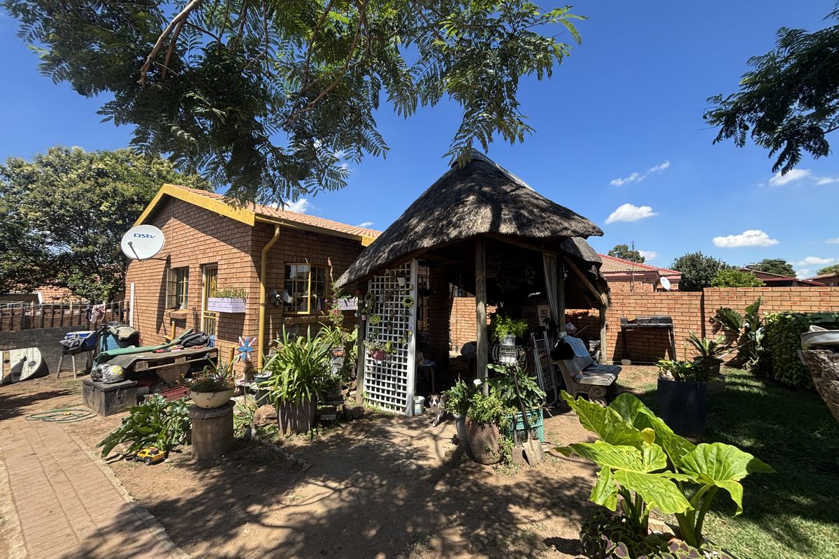 A rear garden area featuring a traditional thatched lapa structure, brick exterior walls, and established landscaping.