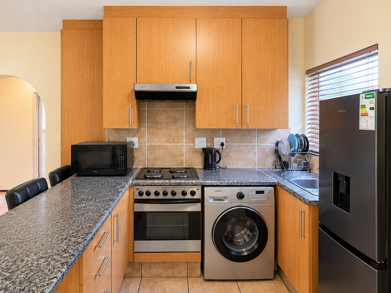 A functional kitchen featuring light wood cabinetry, granite countertops, a tiled backsplash, and integrated appliances including an oven, hob, and washing machine.