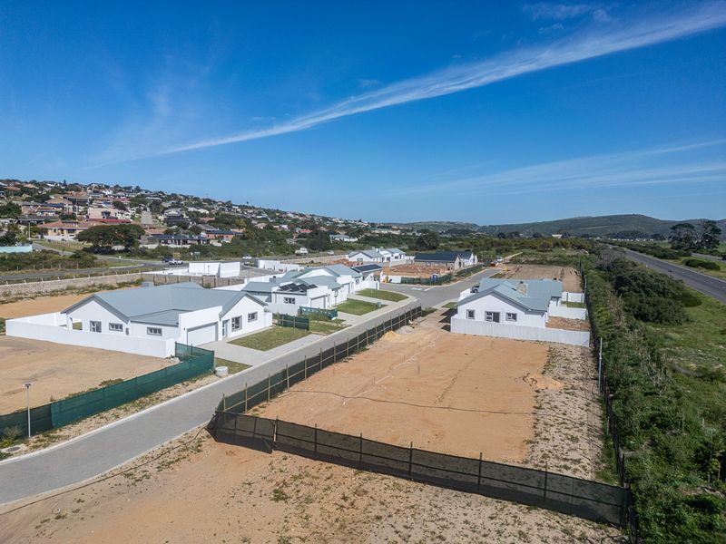 Aerial view of a residential development featuring modern single-story homes with metal roofs, paved driveways, and surrounding undeveloped plots.