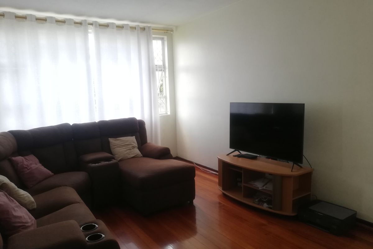 A bright living room featuring polished wooden flooring and large windows with curtains, providing a comfortable and functional space.