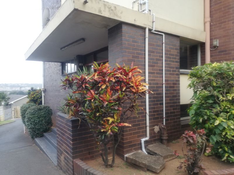 Exterior view of the building facade featuring dark face brickwork, a concrete overhang, and established garden landscaping at the entrance.