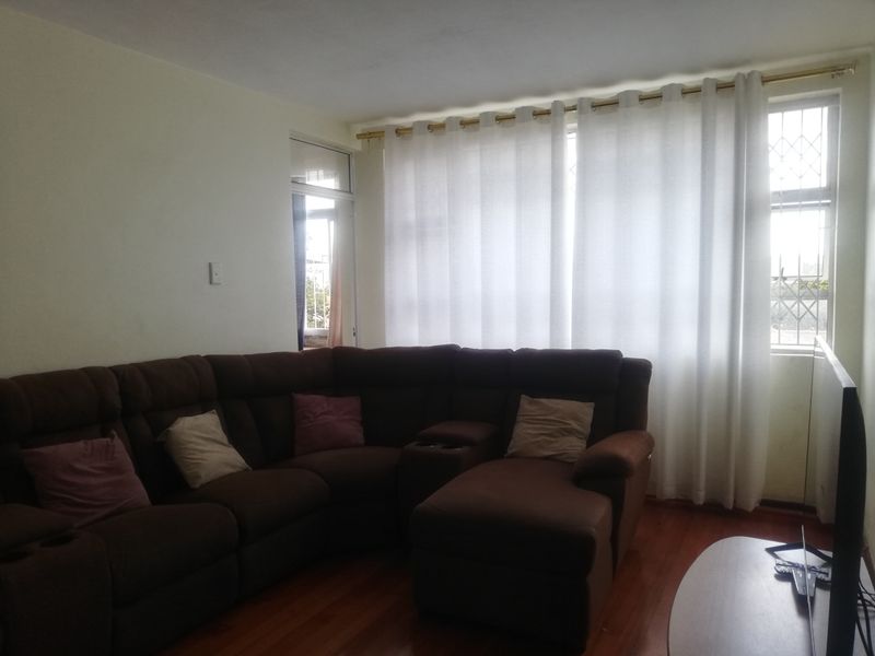 A comfortable living room featuring warm-toned wooden flooring, large windows with privacy curtains, and security bars on the window frames.