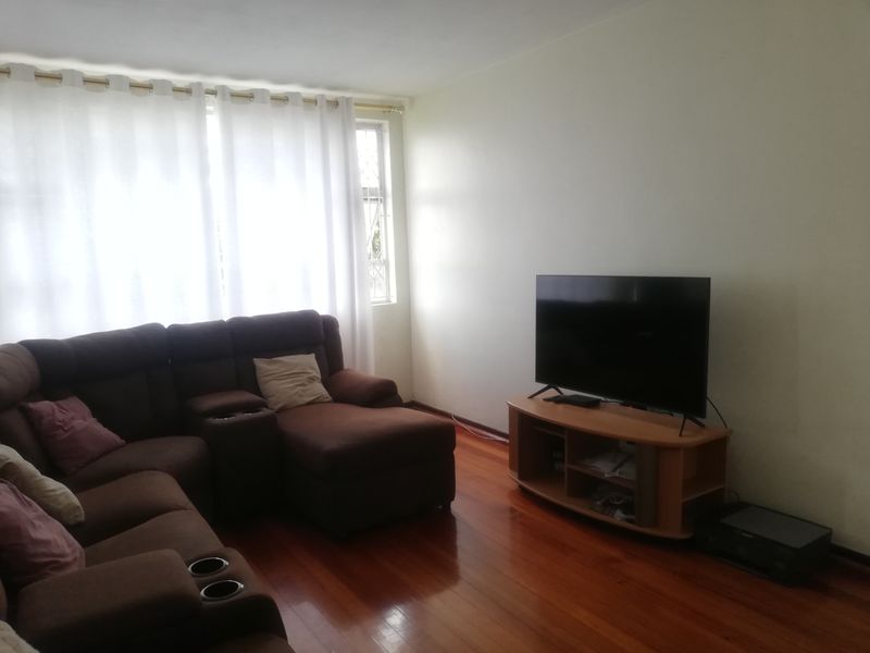 A bright living room featuring polished wooden flooring and large windows with curtains, providing a comfortable and functional space.