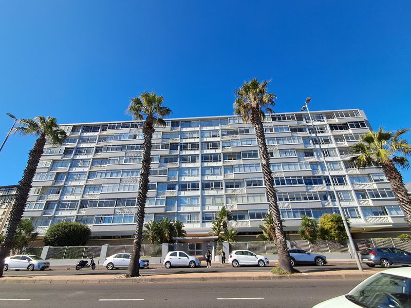 A clear street-level view of a multi-story apartment building facade featuring rows of balconies and established palm trees along the front perimeter.