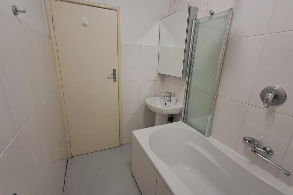 A functional bathroom featuring a pedestal basin, bathtub with a glass shower screen, and wall-mounted mirror cabinet, finished with neutral wall tiles.