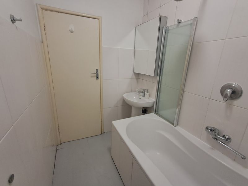 A functional bathroom featuring a pedestal basin, bathtub with a glass shower screen, and wall-mounted mirror cabinet, finished with neutral wall tiles.