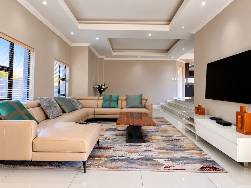 A modern, well-maintained living room featuring a recessed ceiling with downlights, large windows with venetian blinds, and polished tiled flooring.