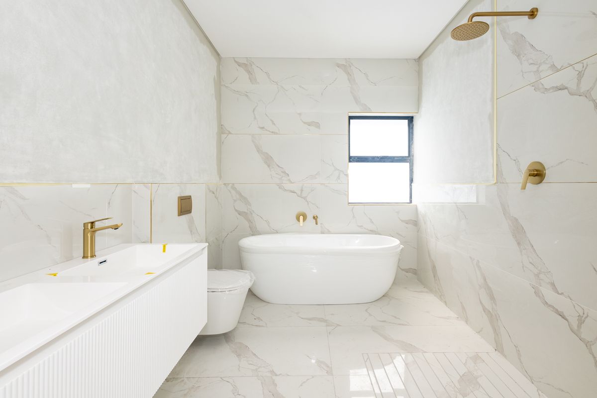 Modern, newly renovated bathroom featuring marble-look tiling, a sleek wall-mounted double vanity, a freestanding bathtub, and elegant brass fixtures.