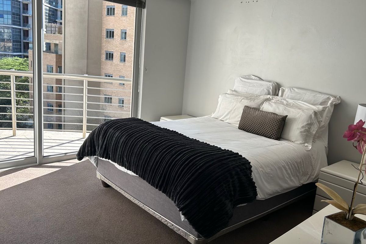 A bedroom featuring carpeted flooring and a large sliding glass door providing access to a private balcony with urban views.