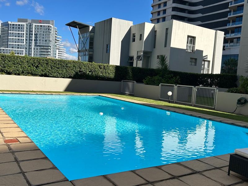 A well-maintained outdoor swimming pool area featuring a paved deck, boundary wall, and landscaped hedging within a modern apartment complex setting.