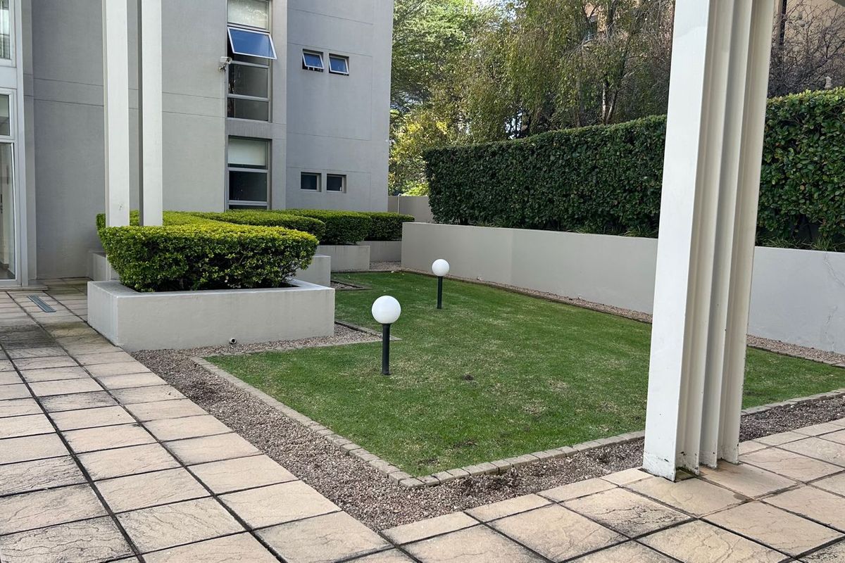 A well-maintained rear courtyard featuring a paved patio area, a small manicured lawn, garden lighting, and established perimeter hedging.