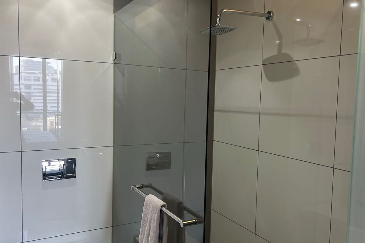 Modern bathroom featuring a walk-in shower with a glass screen, rainfall shower head, and sleek wall-mounted flush plate with neutral ceramic tiling.