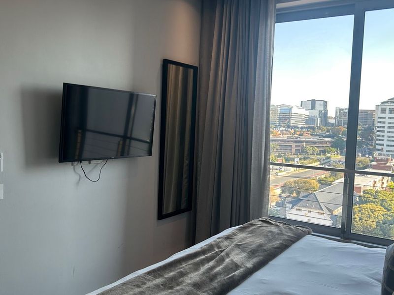 A modern bedroom featuring a large floor-to-ceiling window with city views, neutral wall finishes, and integrated curtain tracks.