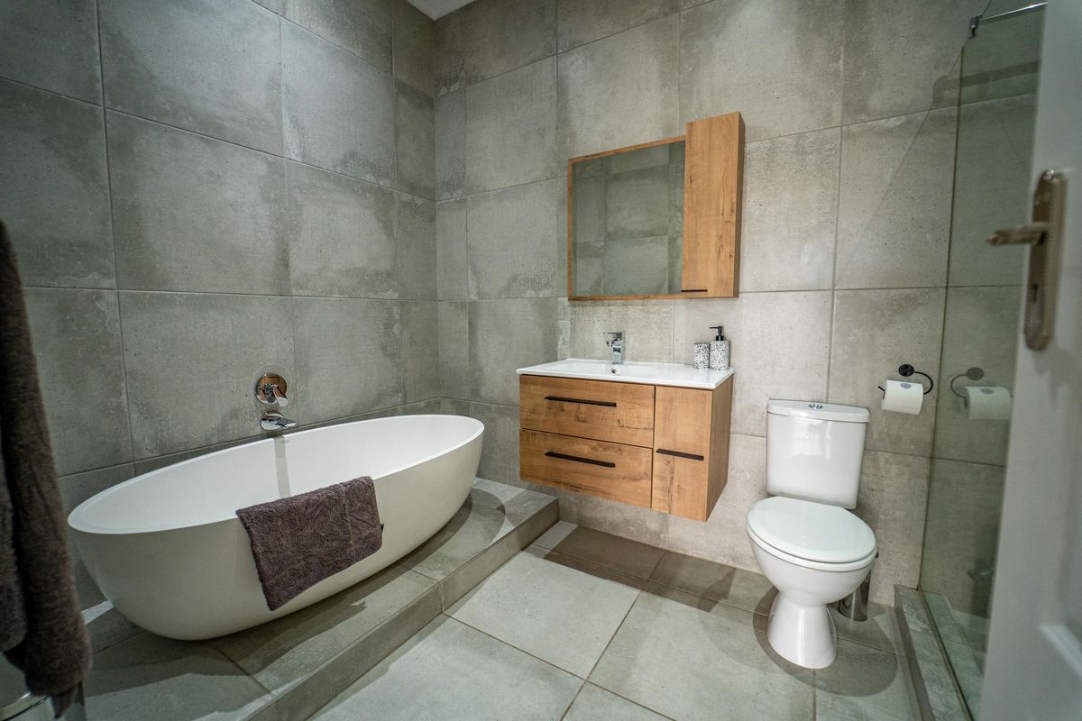 Modern bathroom featuring a freestanding bathtub, wall-mounted wooden vanity with integrated sink, toilet, and a glass-enclosed shower, finished with large format grey tiles.