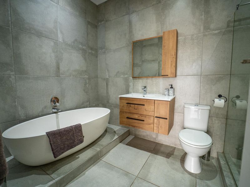 Modern bathroom featuring a freestanding bathtub, wall-mounted wooden vanity with integrated sink, toilet, and a glass-enclosed shower, finished with large format grey tiles.