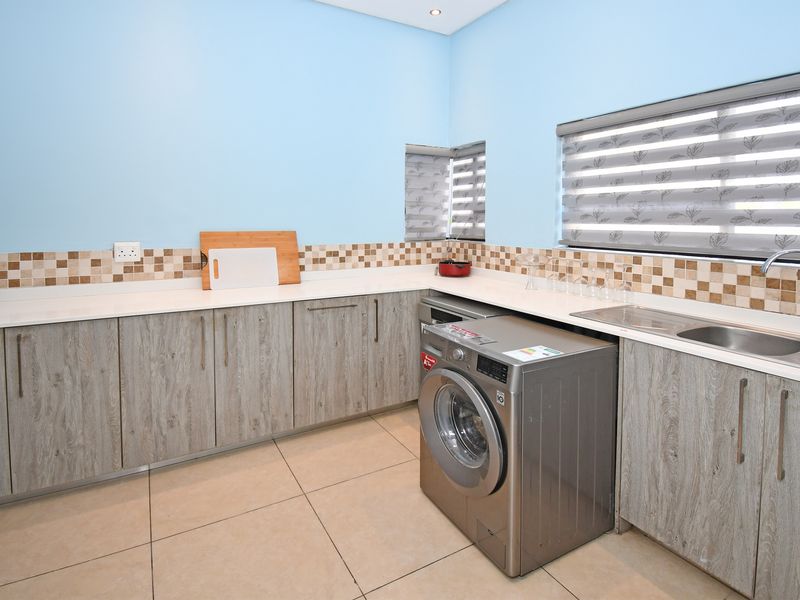 A functional laundry room featuring modern wood-look cabinetry, stone-effect countertops, a mosaic tile backsplash, and a stainless steel double sink with integrated plumbing.