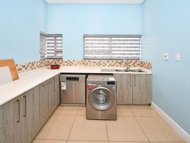 A functional laundry room featuring modern wood-look cabinetry, light countertops, a double sink, and tiled flooring with a neutral backsplash.