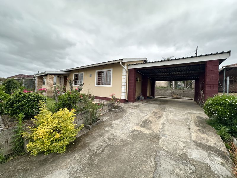 Front view of a single-story house featuring a concrete driveway, attached carport, and established garden beds with security bars on the windows.
