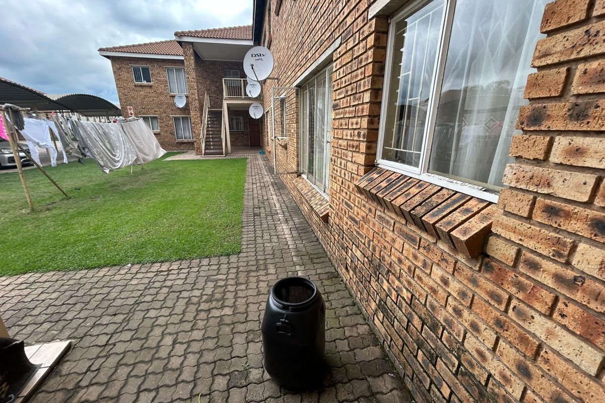 Rear exterior view of a face brick townhouse complex featuring a paved walkway, communal lawn area, and external staircase access.
